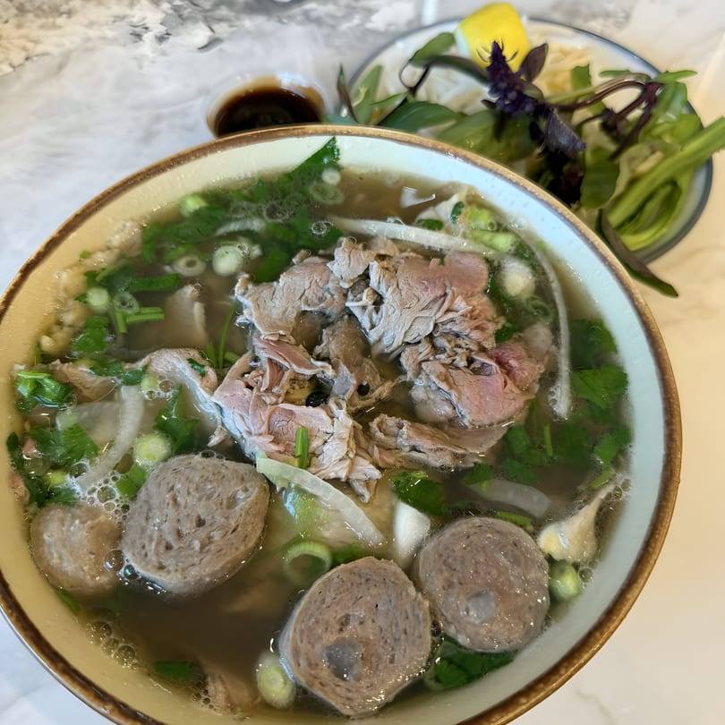 A close-up of a bowl of Vietnamese pho with beef, meatballs, and fresh herbs.