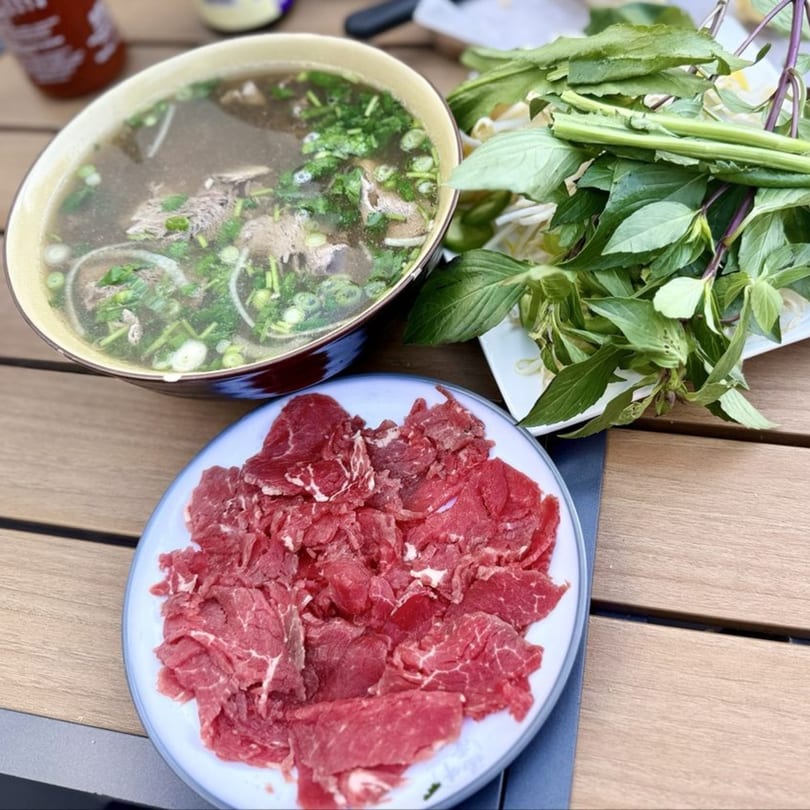 A bowl of PHO 408 pho with thinly sliced beef and fresh herbs on a wooden table.