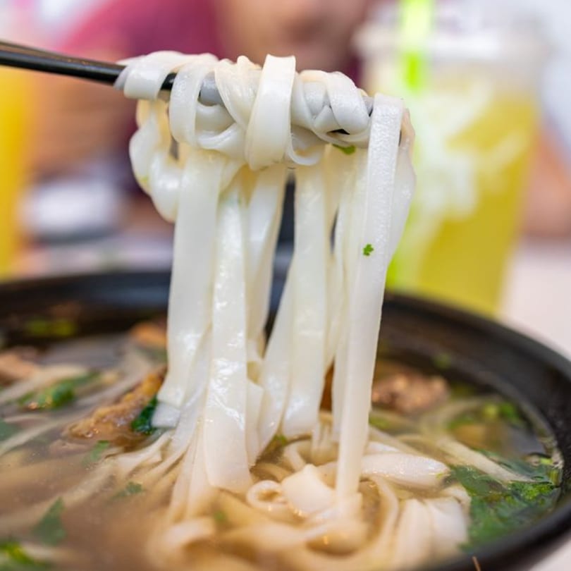 Chopsticks lifting wide rice noodles from a bowl of Vietnamese pho soup.